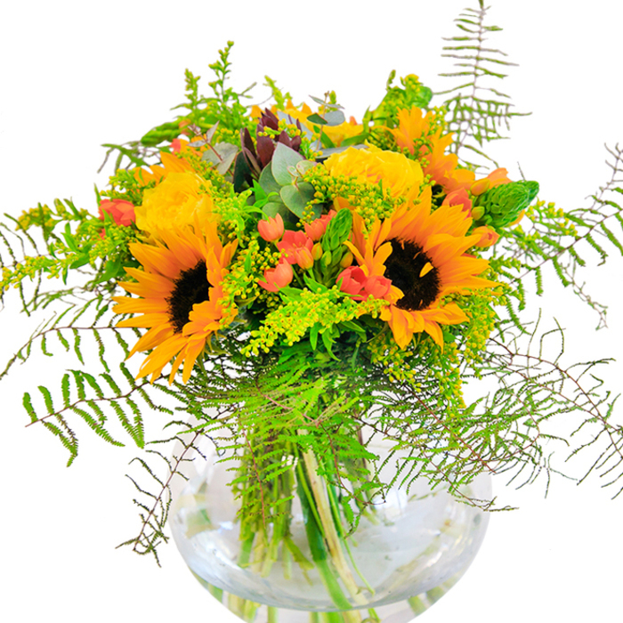 On a crisp North Finchley morning, this sun-inspired bouquet brings the feel of local meadows indoors, expertly gathered and arranged in a pristine glass vase. Sunflowers take centre stage, each bloom radiant with golden petals that radiate around deep, velvety brown centers, evoking the kind of summer brilliance that spills across Finchley Central's green spaces. The arrangement is enhanced by yellow garden roses, their layered, spiralling petals offering a soft, luxurious contrast in both form and scent. Clusters of glossy, red Hypericum berries dot the bouquet like drops of joy, their vibrant hue perfectly offsetting the abundant greenery and lending a playful touch to the composition. Feathery fern fronds drape gently downward, their filigree edges creating an airy, cascading effect that adds both volume and grace. Among the foliage, subtle blue-grey eucalyptus and light green filler sprigs-perhaps goldenrod-peek through, their textures weaving together a tapestry of freshness. The stems, visible in the crystal-clear vase, reveal the care taken in arranging each element; submerged in water, they contribute to an overall impression of vitality and clarity. Crafted by North Finchley Florist, this arrangement is a testament to thoughtful floral artistry. It's well-suited for birthdays, housewarmings, or simply adding a burst of local summer colour to a room near Finchley's bustling markets-an authentic celebration of joy, light, and natural abundance in the heart of North London.